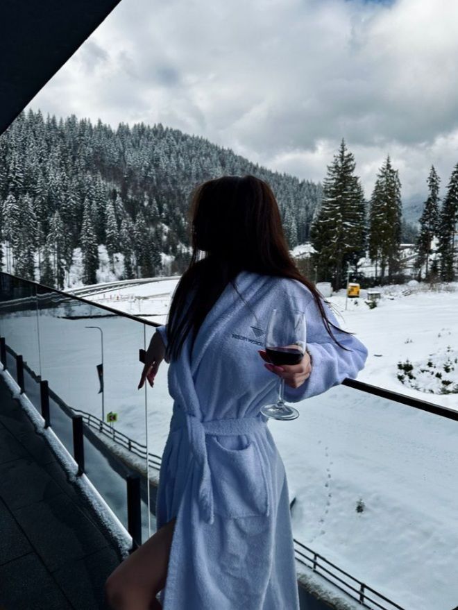 Woman in a robe enjoying a balcony view of a snowy, forested landscape with a glass of wine in hand, exuding relaxation.