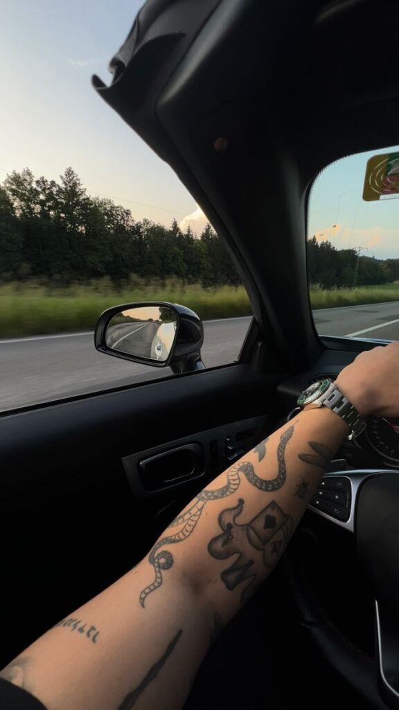 Tattooed arm steering a car, with scenic highway view, lush trees, and sunset sky out the window. Road trip adventure.