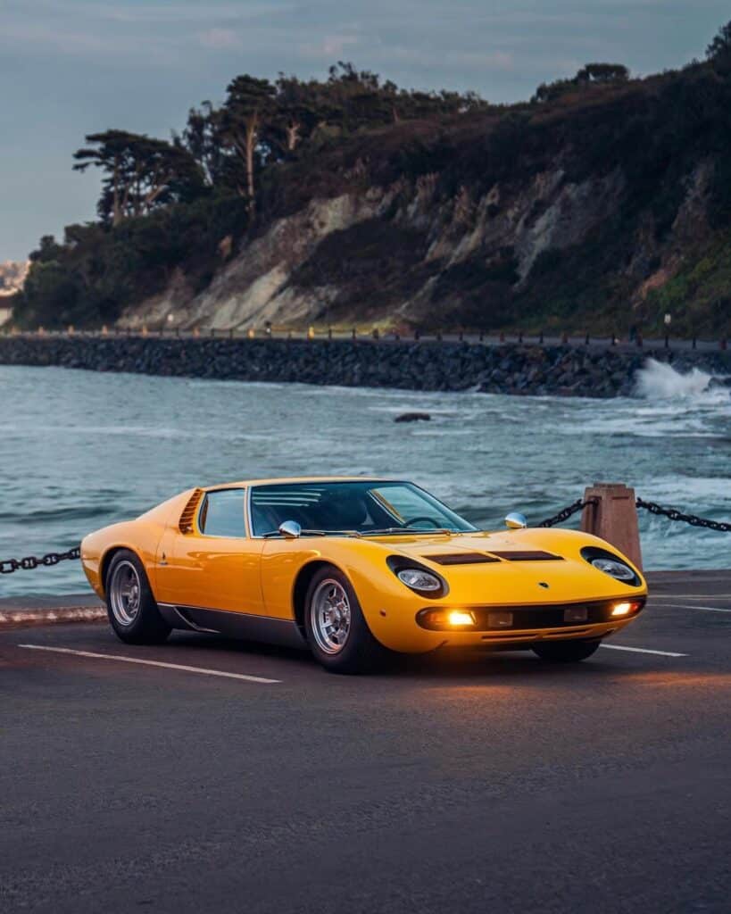 9f26288f39180effc74845a0502109bf Yellow classic sports car parked by the ocean with coastal cliffs and trees in the background at sunset. | Sky Rye Design Yellow classic sports car parked by the ocean with coastal cliffs and trees in the background at sunset.