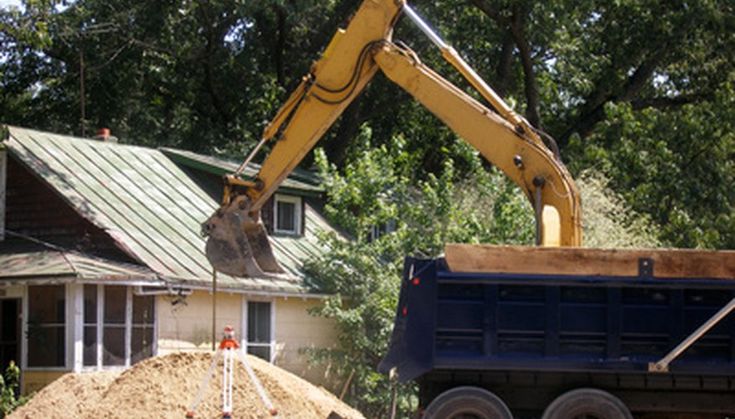 Excavator loading sand into a dump truck at a construction site near a house with green roofing, surrounded by trees.