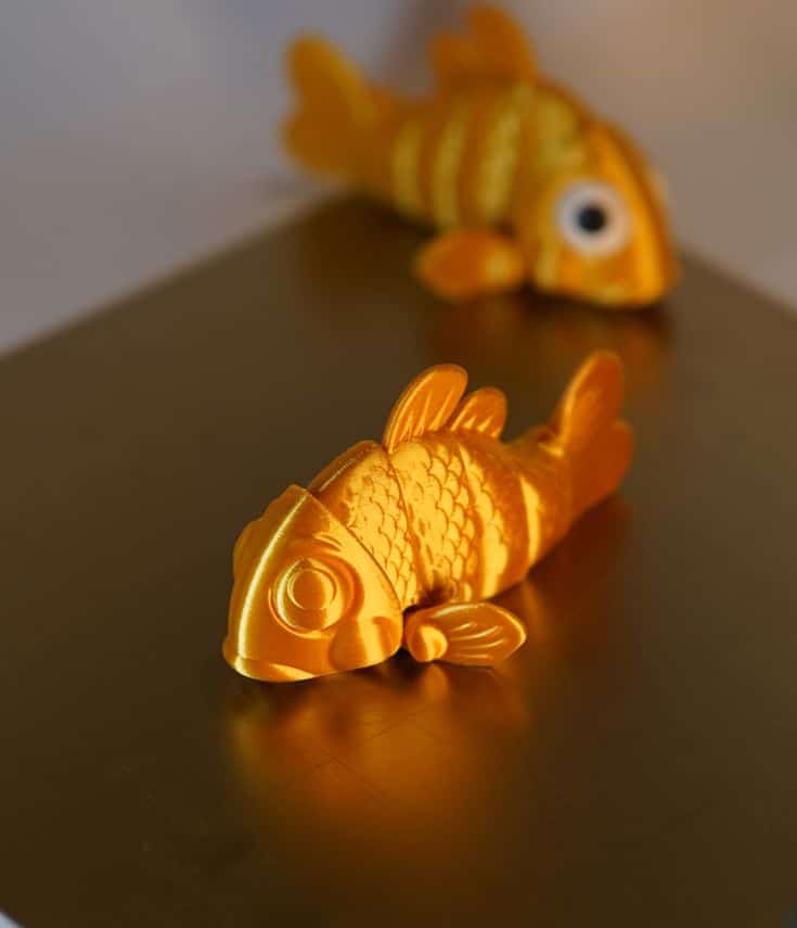 Golden 3D-printed fish model with intricate scales, set against a warm-toned background, while another fish blurs in the distance.