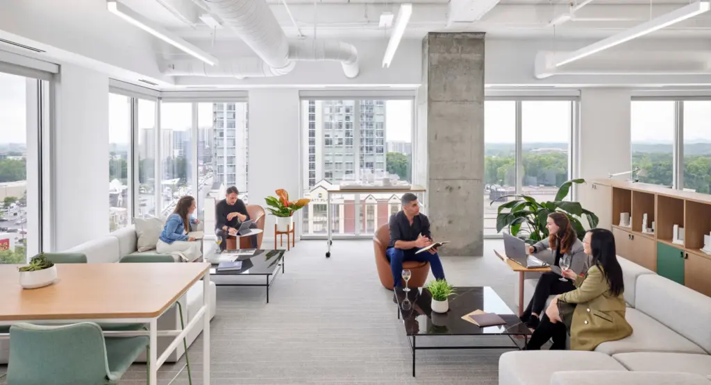 Modern office lounge with people collaborating, featuring large windows, plants, and city views, creating a bright and open workspace.