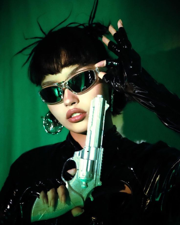 Futuristic fashion: person in sleek black attire, wearing reflective sunglasses, holding a silver toy gun, under green lighting.