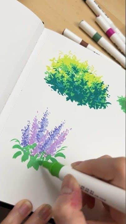 Hand drawing colorful flowers in a sketchbook with markers, featuring lavender blossoms and green foliage.