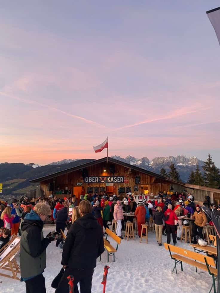 Crowded mountain hut with skiers enjoying après-ski under a pink sunset sky in the snowy Alps, flag waving overhead.