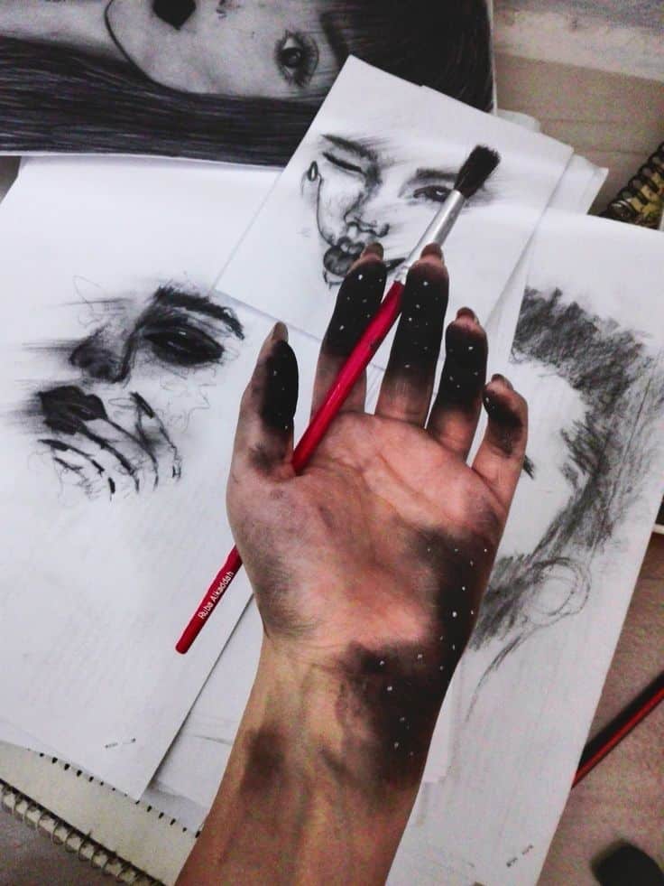 Artist's hand covered in charcoal, holding a brush, with expressive sketches in the background, showcasing creative process.