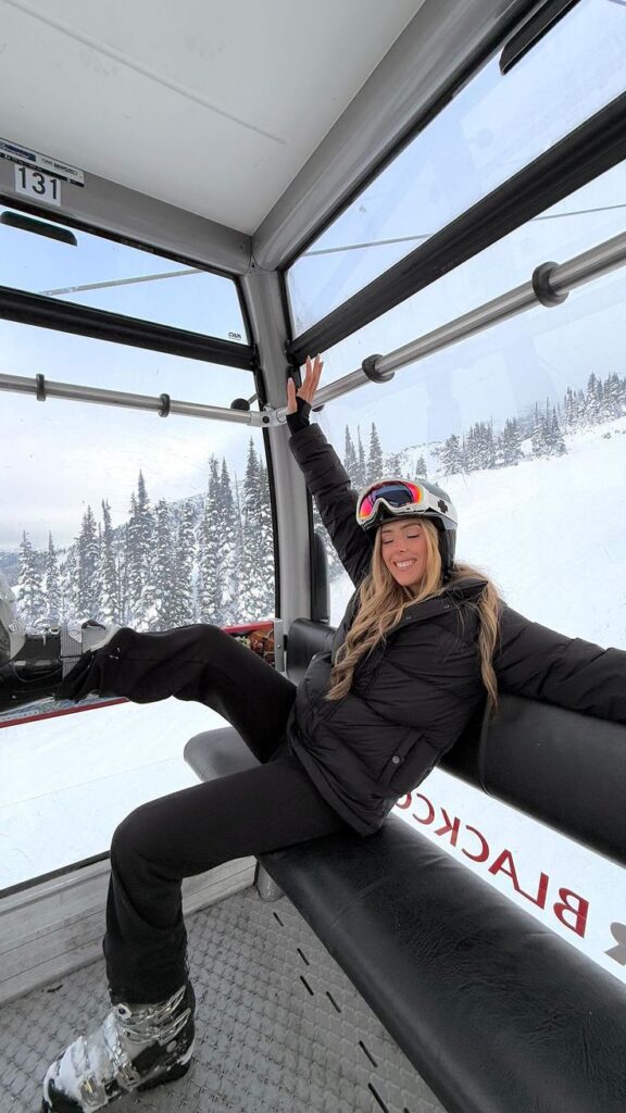 Person smiling and relaxing on a snowy gondola ride in a winter landscape, wearing ski gear with snow-covered trees outside.