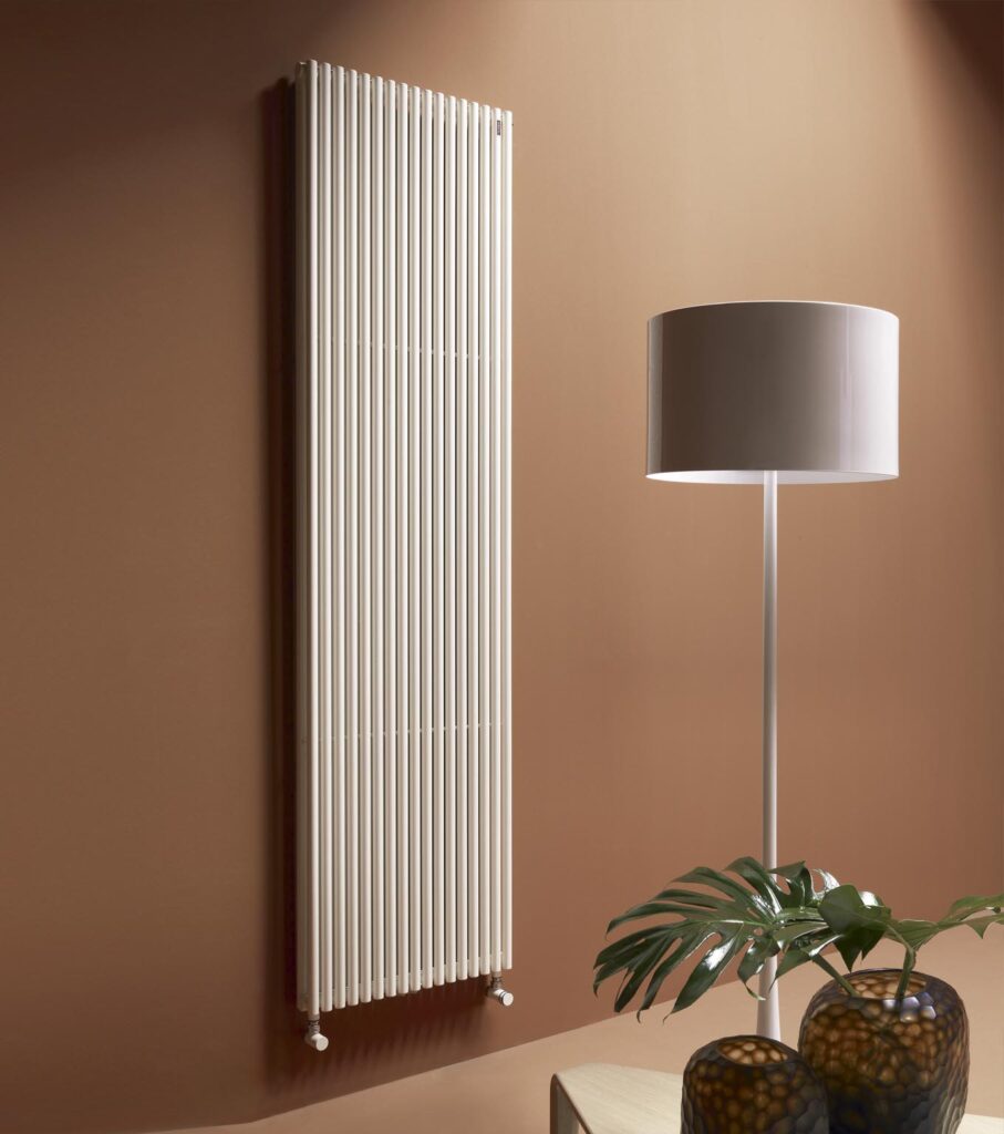 Sleek white vertical radiator on brown wall, beside a modern lamp and decorative vases. Stylish, minimalist home heating solution.