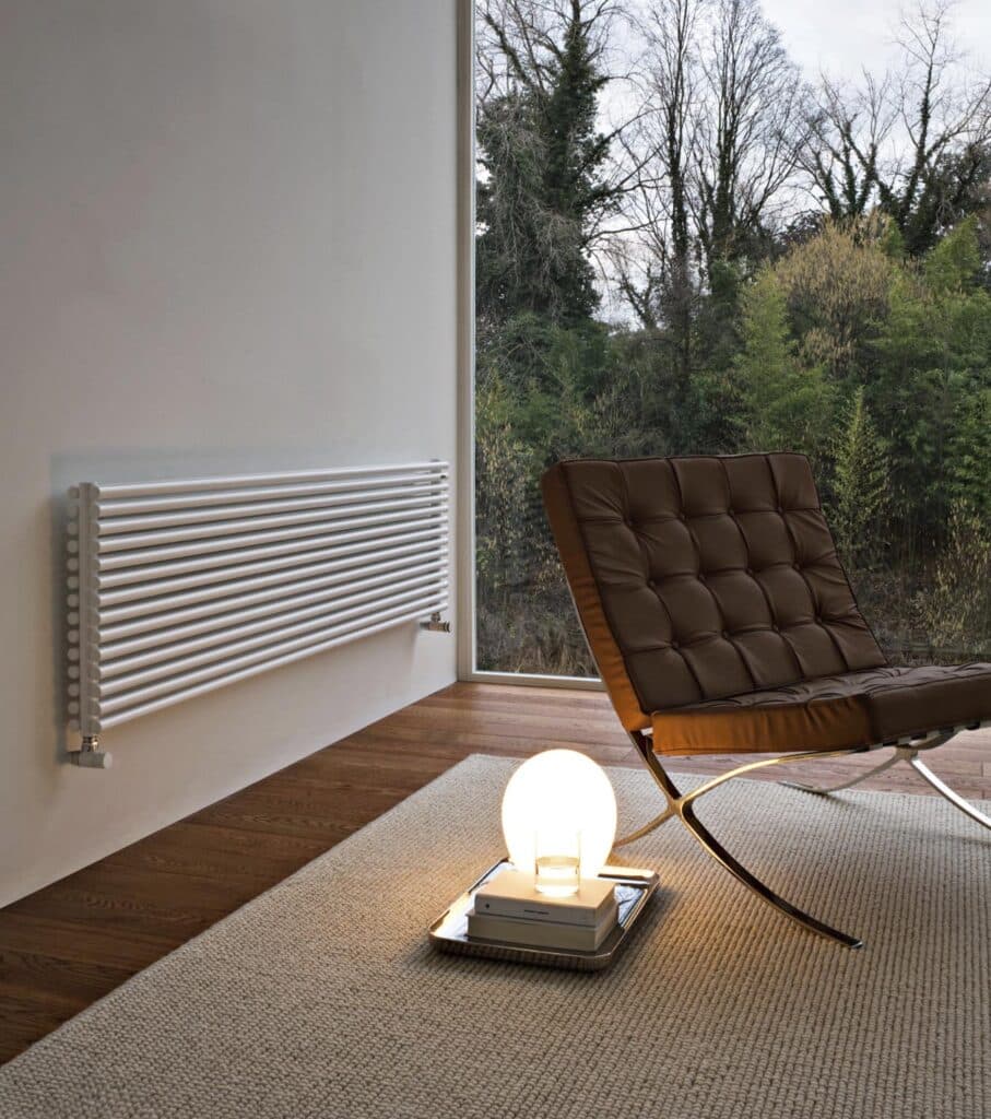 Modern minimalist room with brown lounge chair, white radiator, soft rug, and glowing globe lamp, overlooking lush greenery through a large window.