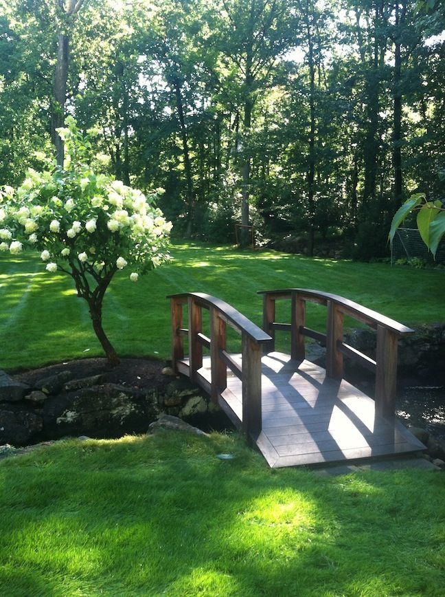 Tranquil garden scene with a wooden bridge over a small stream, surrounded by lush greenery and blooming white flowers in sunlight.