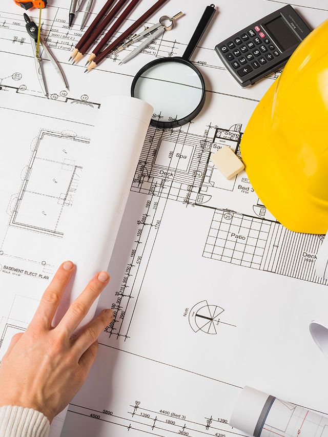Best Contractors in Muscat Oman_ Contracting and Construction Company in Muscat Oman Architectural blueprints with tools, calculator, and hard hat; hand examining design plans on a workspace. | Sky Rye Design Architectural blueprints with tools, calculator, and hard hat; hand examining design plans on a workspace.
