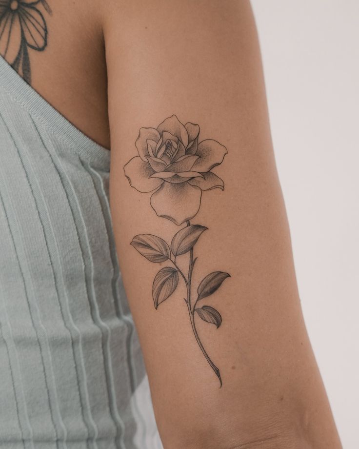 Black and gray rose tattoo on upper arm, showing intricate details of petals and leaves, against a sleeveless top.
