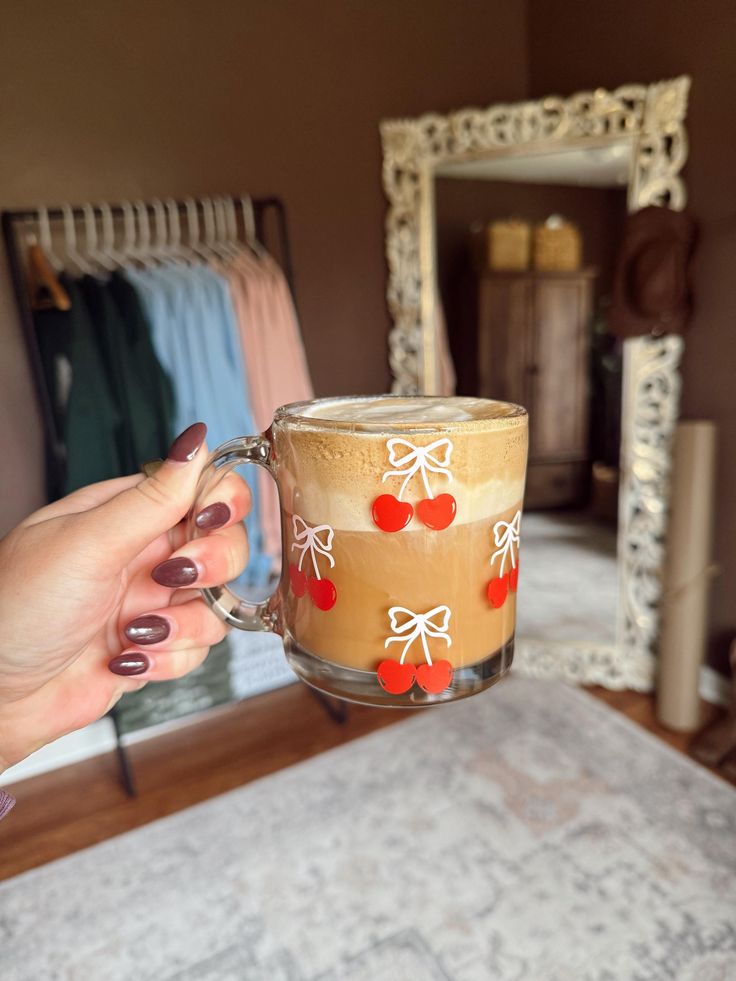 Bow Glass Coffee Cup _ Aesthetic Iced Coffee Glass _ Cute Mug for Her _ Trendy Bow Design _ Gift for Coffee Lovers Hand holding a latte in a cherry-themed mug, with a cozy room in the background featuring clothes and a decorative mirror. | Sky Rye Design Hand holding a latte in a cherry-themed mug, with a cozy room in the background featuring clothes and a decorative mirror.