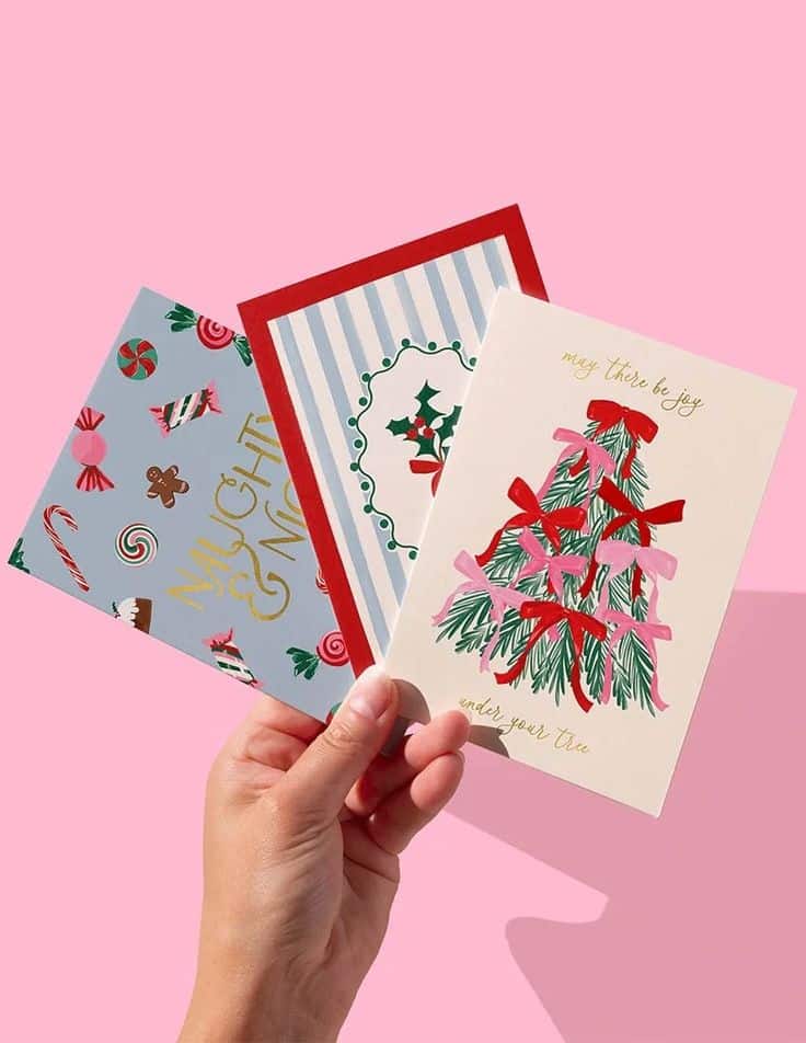 Hand holding festive holiday cards with Christmas designs against a pink background, featuring a tree and holly artwork.