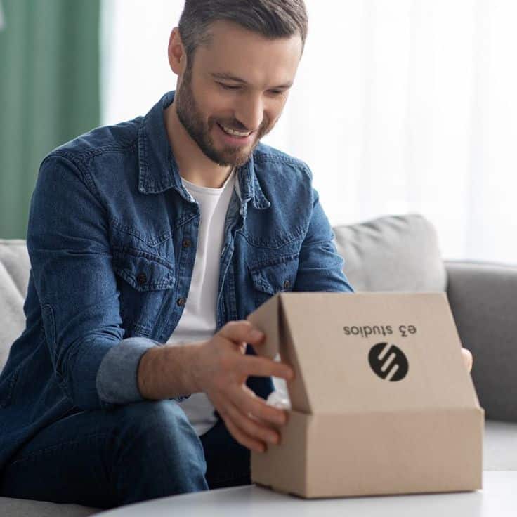 Man in denim shirt happily unboxing a package on the couch, branded with 'e3 studios.'