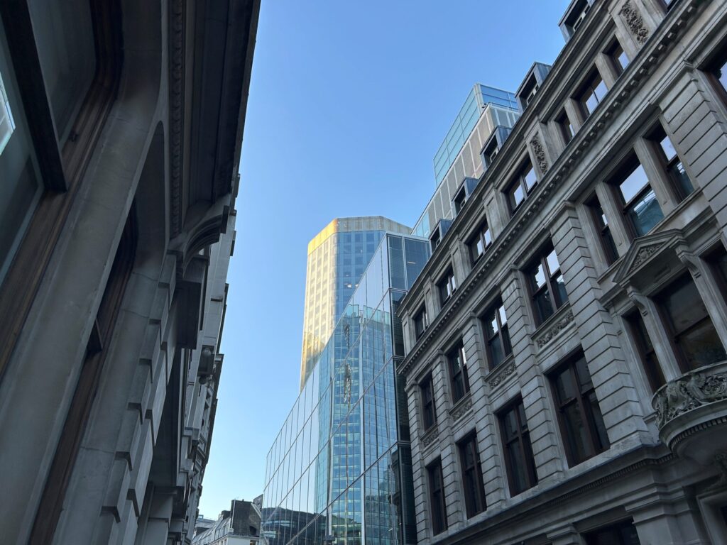 Modern glass skyscraper framed by historic architecture under a clear blue sky. Contrast of old and new in urban cityscape.