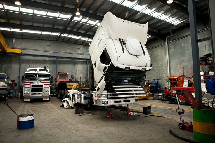 Truck maintenance in a workshop, with an elevated cab for repairs. Heavy-duty equipment and tools visible in the industrial garage.