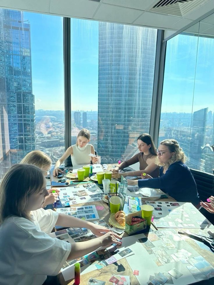 Group of people working on a creative project in a modern office with city views, surrounded by photos and art supplies on the table.