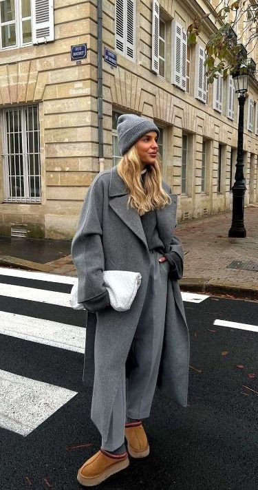 Woman in stylish gray coat and beanie at a crosswalk, holding a white clutch on a city street.