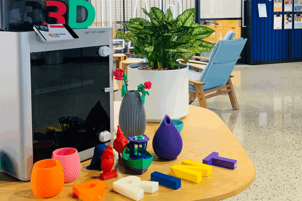 3D printer with colorful printed objects, plants, and modern office decor on a wooden table, showcasing creativity and innovation.