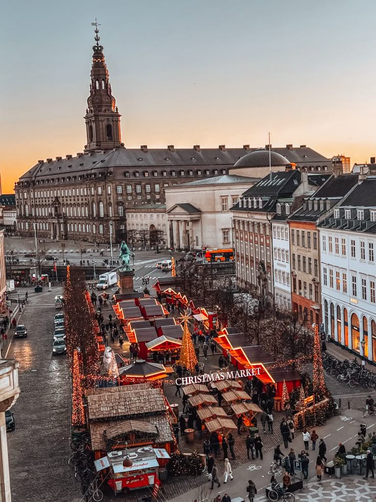 Christmas market in a European city square at sunset, featuring festive lights, busy stalls, and historic architecture.