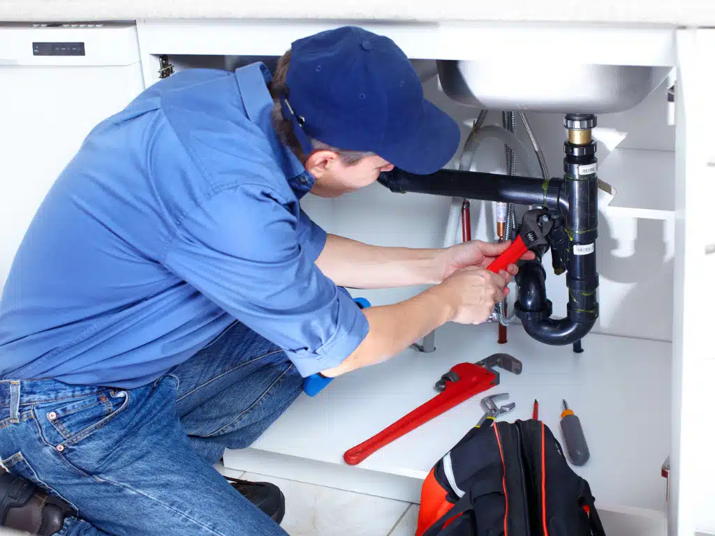Plumber fixing kitchen sink pipes with wrench, wearing blue uniform. Tools visible, ensuring smooth water flow and maintenance.