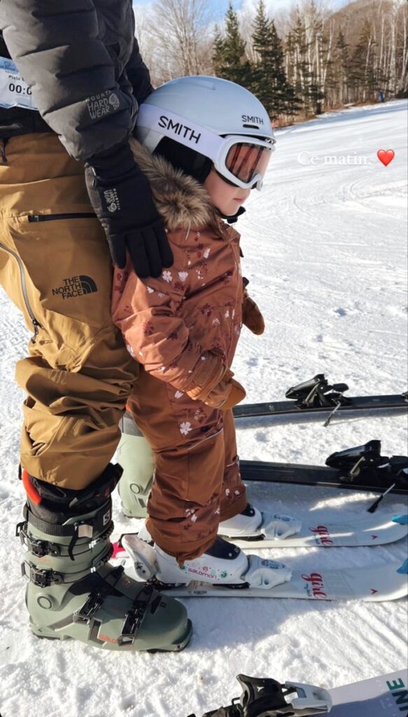 Child learning to ski with assistance on a sunny winter day, wearing snow gear and helmet, surrounded by snow and ski gear.