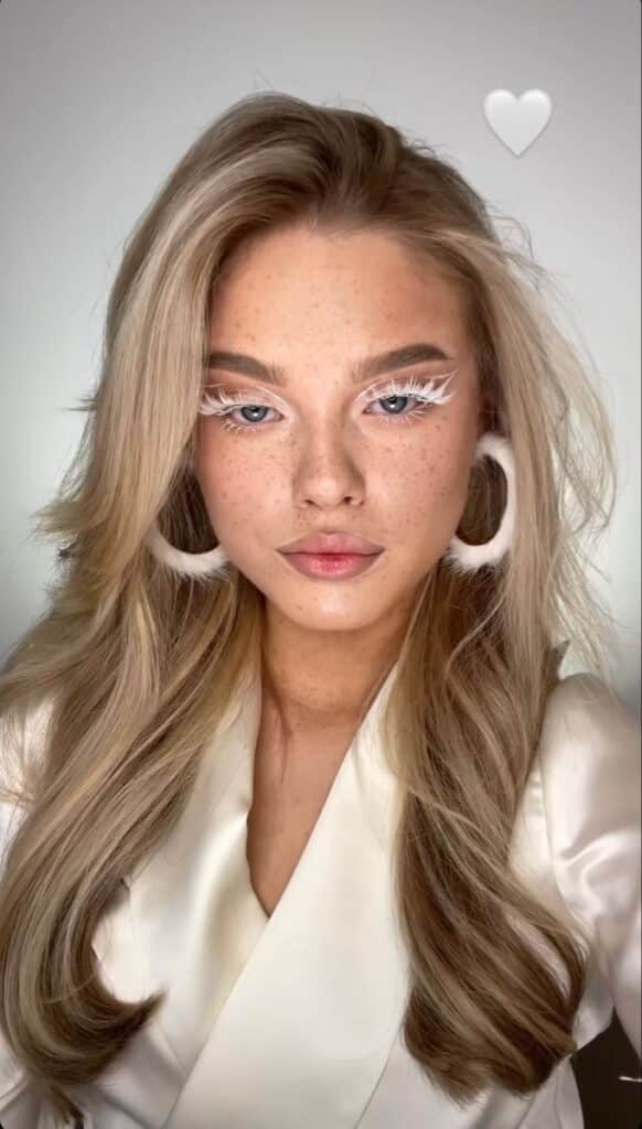 Woman with long blonde hair, white eye makeup, and bold accessories in a stylish, elegant attire, posing with a serene expression.