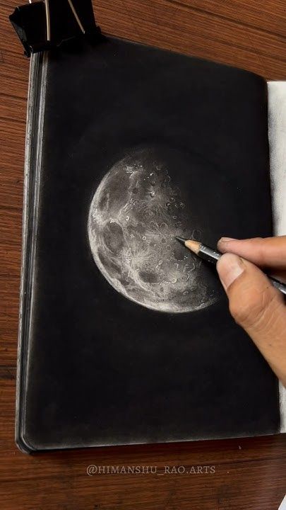 Artist sketching a detailed moon drawing in a notebook using a pencil, highlighting craters and shadows on a wooden desk background.