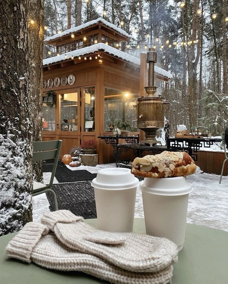 Cozy winter scene at a snowy outdoor cafe with warm drinks, pastries, and mittens on the table, surrounded by twinkling lights in the forest.
