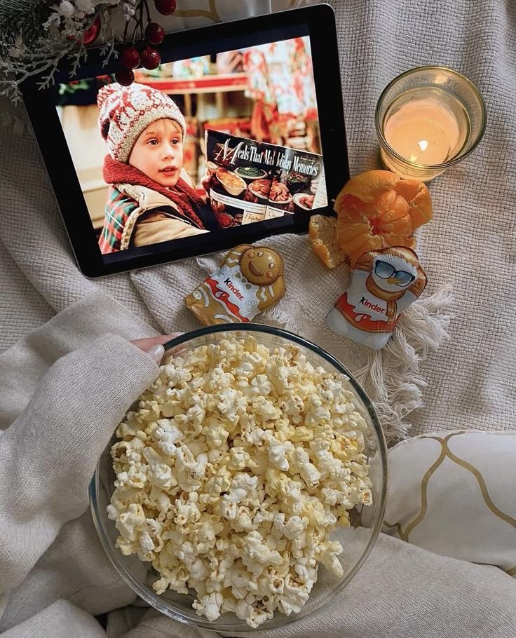 Cozy movie night setup with popcorn, holiday treats, and a lit candle on a tablet screen showing a classic holiday movie.