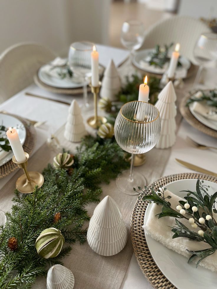 Elegant Christmas table setting with candles, white ceramic trees, greenery, and festive decorations on a woven placemat.
