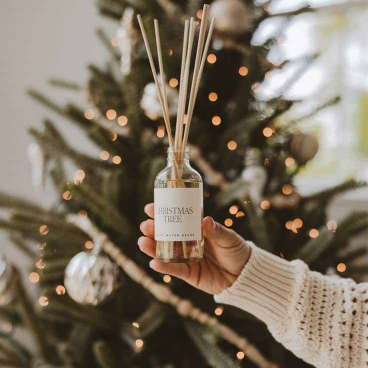 Christmas Tree Clear Reed Diffuser Hand holding a Christmas tree scented diffuser in front of a decorated tree with warm lights and ornaments in a cozy holiday setting. | Sky Rye Design Hand holding a Christmas tree scented diffuser in front of a decorated tree with warm lights and ornaments in a cozy holiday setting.