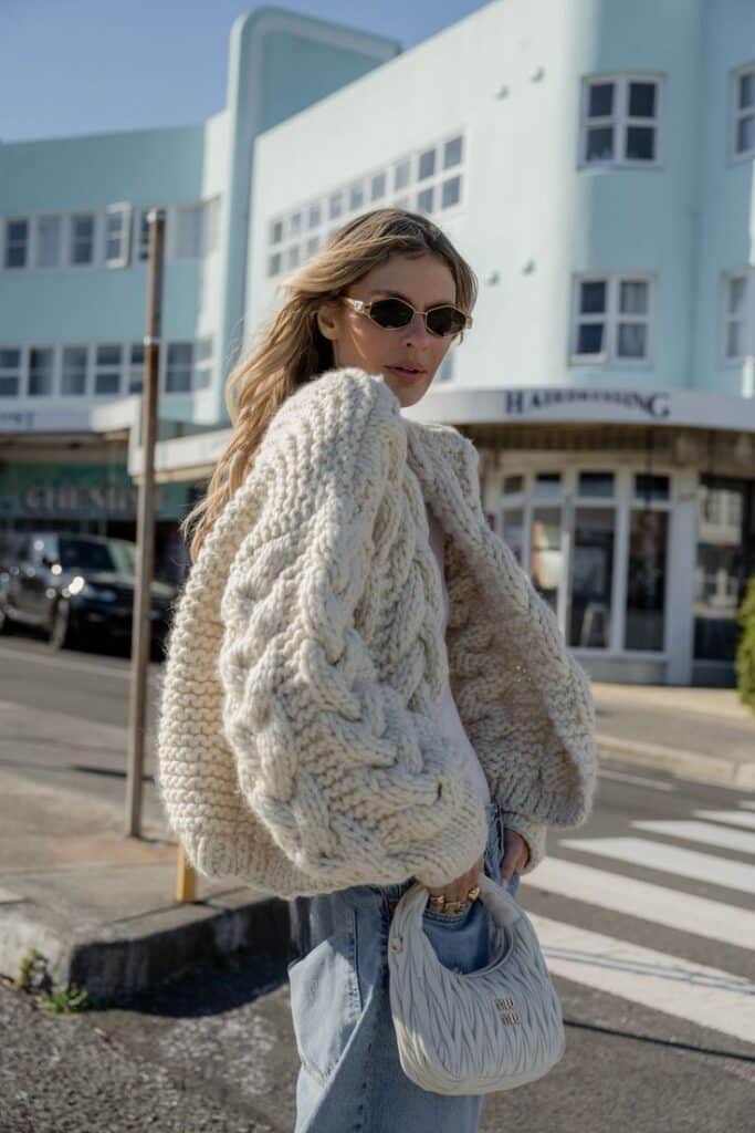 Chunky Cable Cardigan Fashionable woman in chunky knit sweater and sunglasses holds bag, walking in urban setting. | Sky Rye Design Fashionable woman in chunky knit sweater and sunglasses holds bag, walking in urban setting.