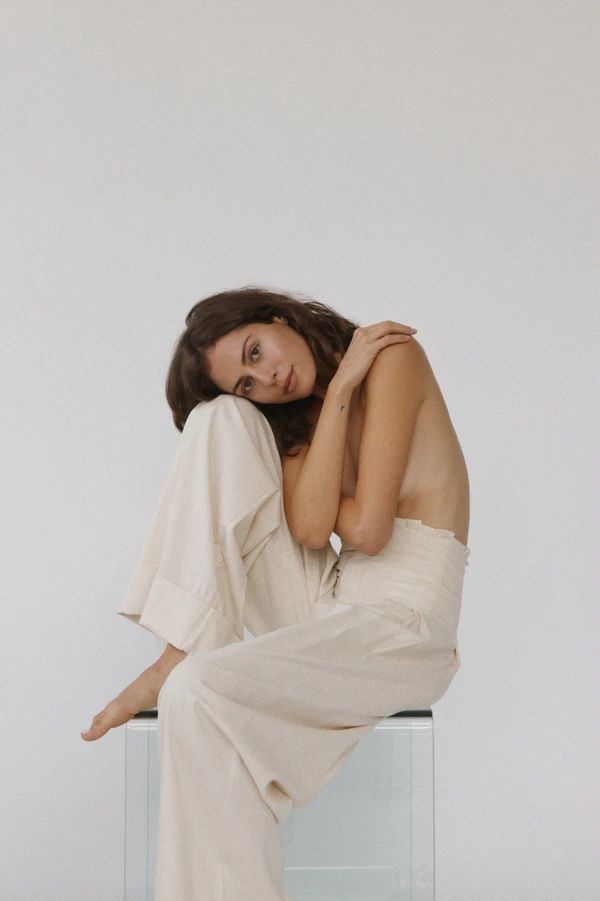 Woman in light pants poses seated on a clear box, embracing knees, with soft light and minimalist background.
