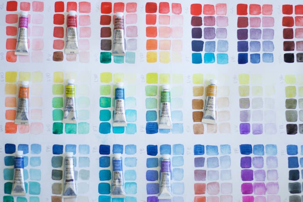 Assorted watercolor tubes on a grid of vibrant color swatches, showcasing various shades from reds to blues, ideal for artists and designers.