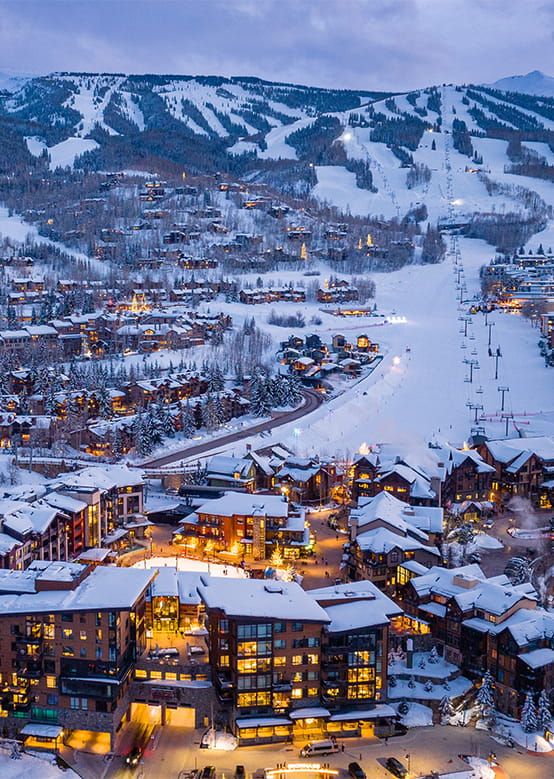 Snow-covered mountain village at dusk with ski slopes and illuminated buildings.