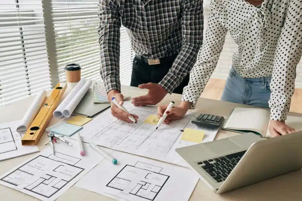 Construction-Estimating-Services-1 Architects discussing floor plans at a desk with blueprints, laptop, and design tools. | Sky Rye Design Architects discussing floor plans at a desk with blueprints, laptop, and design tools.