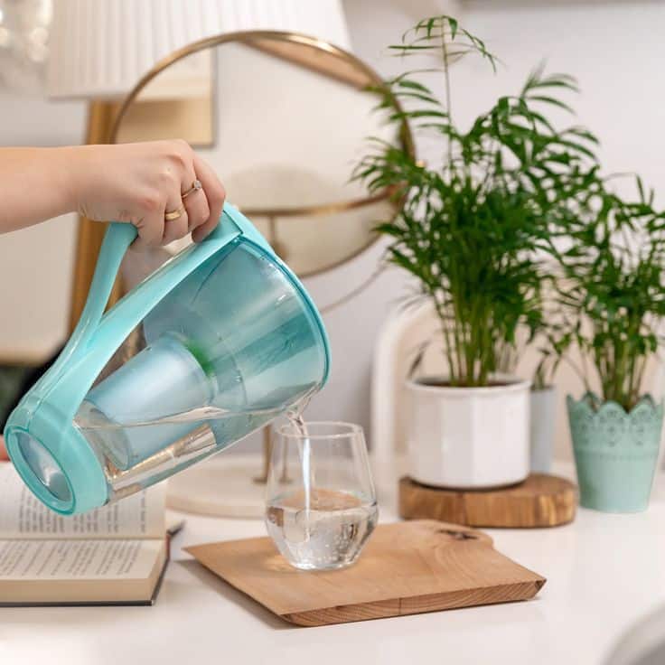 Crystal Water Filter Pitcher Person pouring water from a blue filter jug into a glass on a table with indoor plants and books. | Sky Rye Design Person pouring water from a blue filter jug into a glass on a table with indoor plants and books.