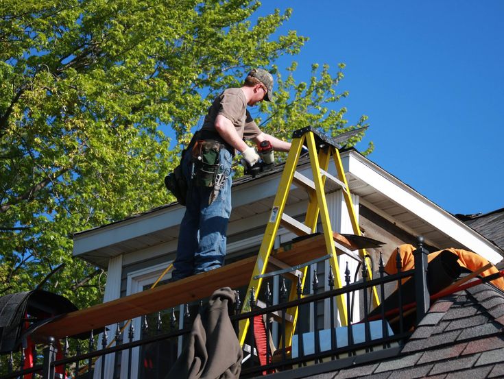 Roof technician working on a house, using a ladder and tools, under a clear blue sky with green trees in the background.