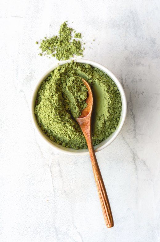 Bowl of vibrant green matcha powder with wooden spoon on a light marble surface. Perfect for tea or recipes.