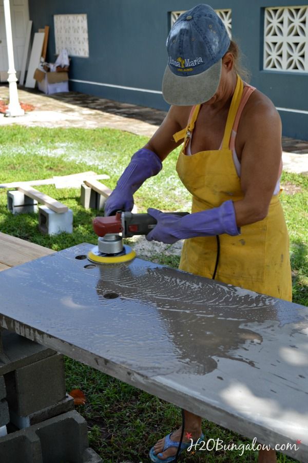 Person polishing a concrete countertop outdoors, wearing a cap, yellow apron, and purple gloves. Grass and building wall in background.