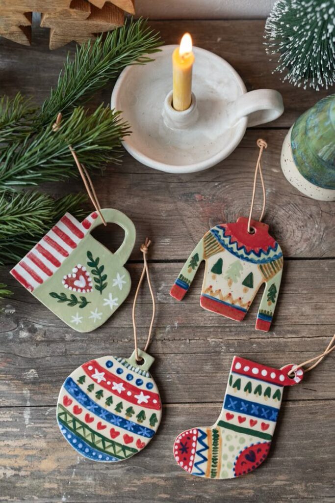 Festive handcrafted Christmas ornaments, including a sweater, mug, stocking, and bauble, next to a candle on a rustic wooden table.