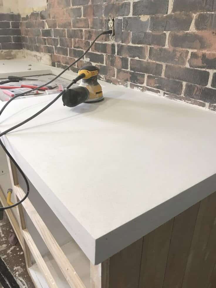 Electric sander on white countertop with exposed brick wall background in a home renovation scene.