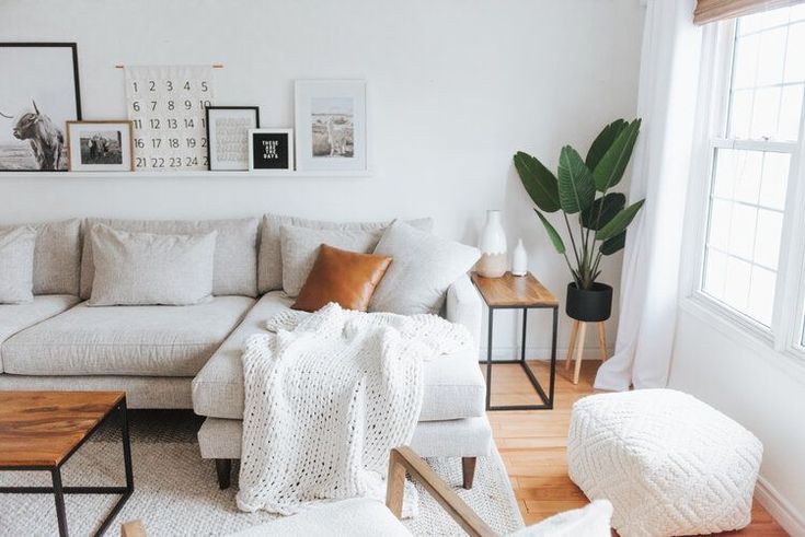 Modern living room with a cozy beige sectional sofa, knit blanket, and houseplant by the window, featuring minimalist wall art and wooden accents.