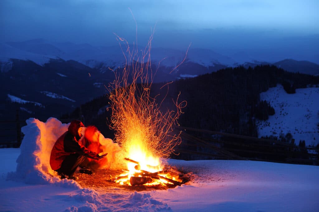 Person warming by a campfire in snowy mountains at dusk, with sparks flying into the cold night sky.