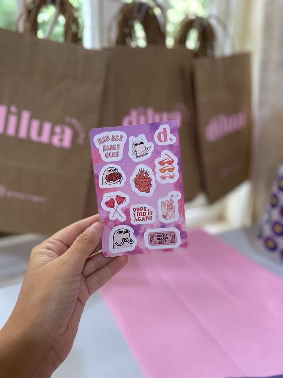 Dicas de Brindes para Clientes _ Loja Online ♡ Hand holding a purple sheet of quirky stickers with text and doodles, against a background of brown shopping bags with pink branding. | Sky Rye Design Hand holding a purple sheet of quirky stickers with text and doodles, against a background of brown shopping bags with pink branding.