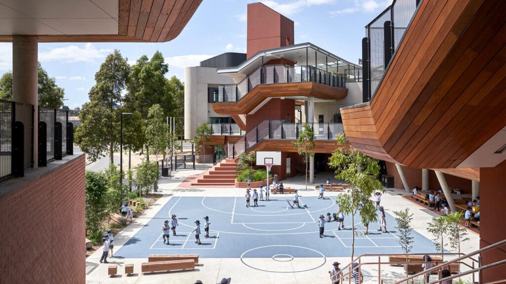 Docklands-Primary-School-Photo-5 School courtyard with modern architecture, featuring students playing on a blue basketball court surrounded by trees and benches. | Sky Rye Design School courtyard with modern architecture, featuring students playing on a blue basketball court surrounded by trees and benches.