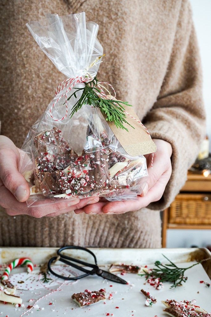 Easy Candy Cane Chocolate Bark 3 Ingredients No-Bake Person holding a festive bag of holiday chocolate bark, decorated with peppermint and tied with a twine and pine sprig. | Sky Rye Design Person holding a festive bag of holiday chocolate bark, decorated with peppermint and tied with a twine and pine sprig.