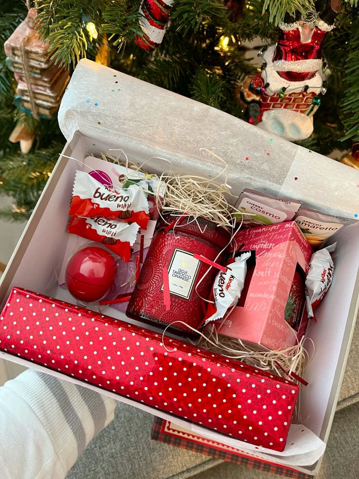 Easy Christmas gift for everyone Gift box with red candle, chocolates, and beauty items nestled in festive holiday packaging, near a decorated Christmas tree. | Sky Rye Design Gift box with red candle, chocolates, and beauty items nestled in festive holiday packaging, near a decorated Christmas tree.